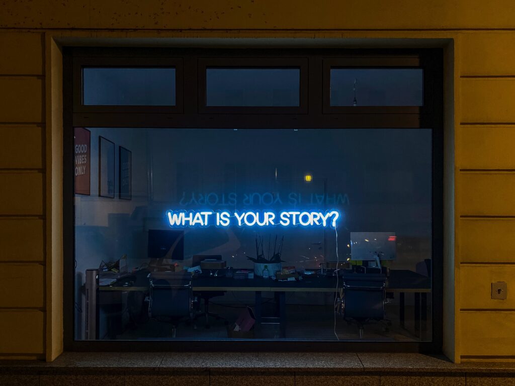 a neon sign reading "what is your story" hangs in a darkened window